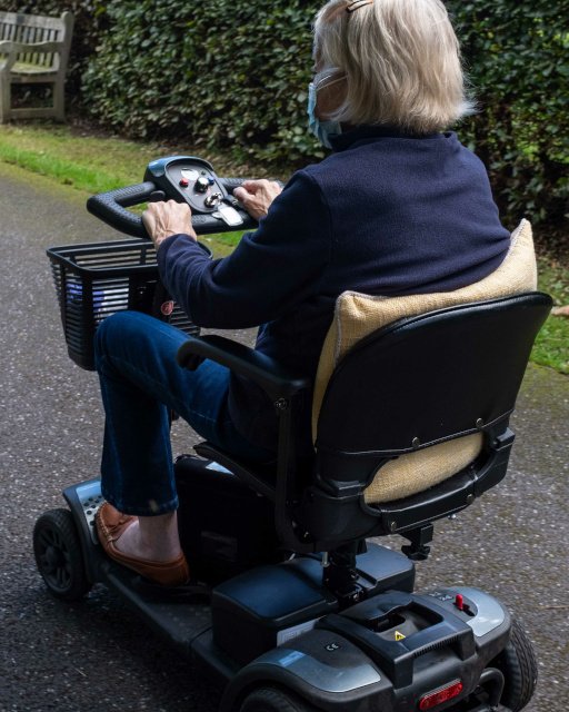 An old lady in a blue jacket riding a mobility scooter on a quiet lane from behind three quarters view